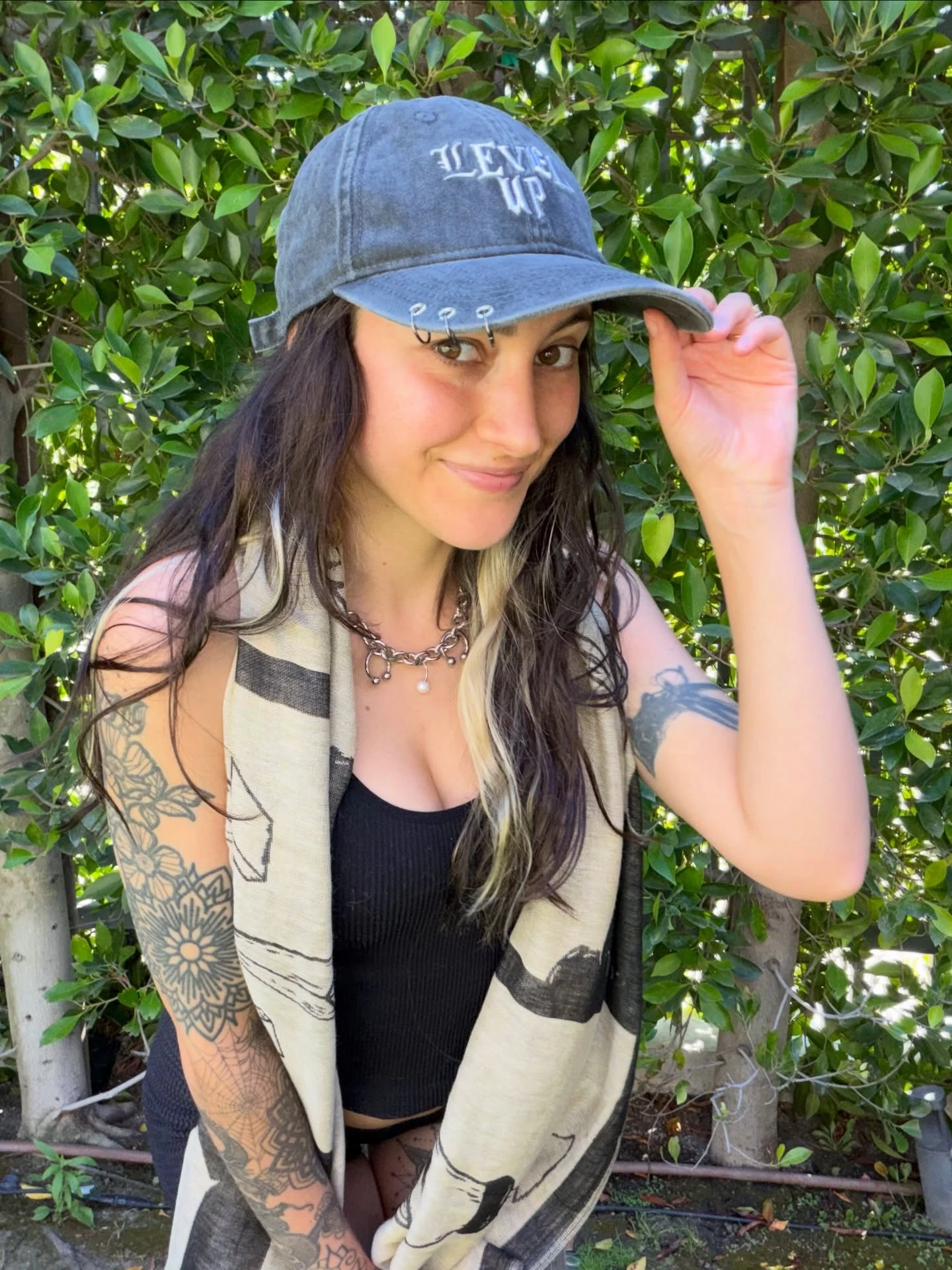 Level Up tipping a washed gray Level Up dad hat with pierced rings on the brim, wearing a bone-print scarf in front of a leafy hedge